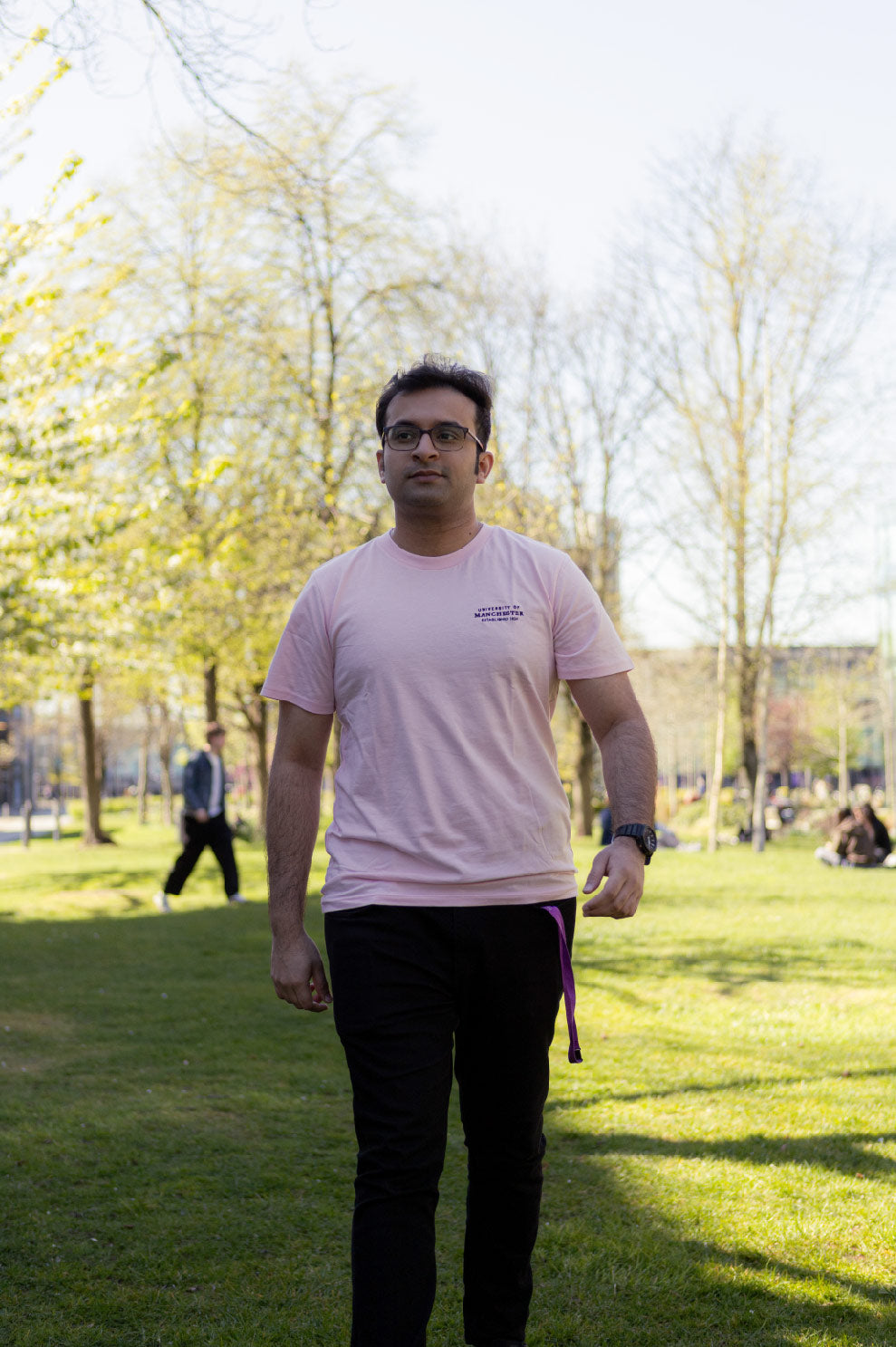 A person in a pink T-shirt and black pants walks confidently through a sunlit park, with trees and people in the background, conveying a relaxed atmosphere.