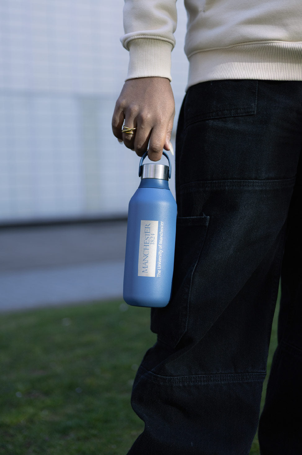 A person in a cream sweater and black pants holds a blue water bottle with "University of Manchester" branding. The scene conveys a casual and relaxed outdoor setting.