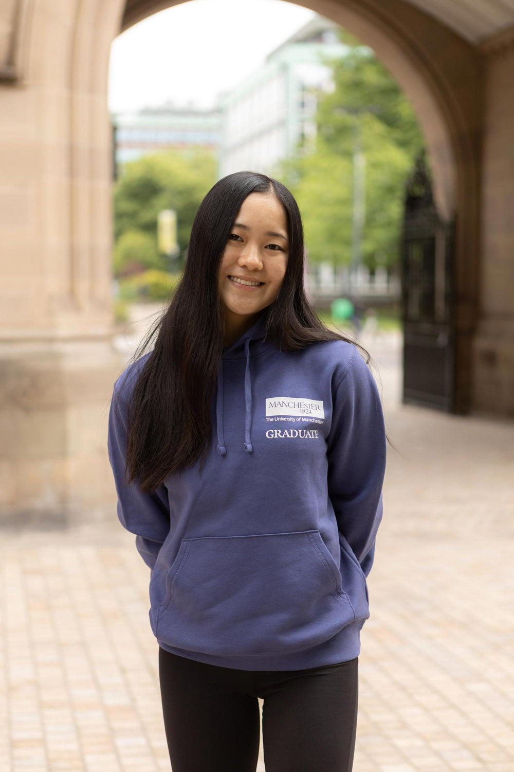 Smiling person wearing a purple University of Manchester Graduate hoodie stands under an archway. Background includes trees and modern buildings.