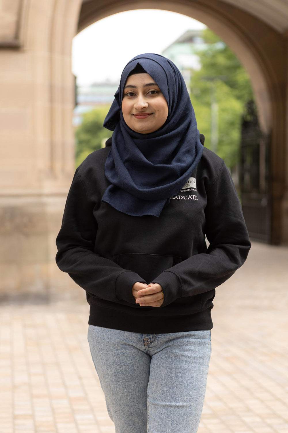 Person in a black "University of Manchester Graduate" hoodie smiles confidently. She's outdoors, framed by an archway with trees in the background.