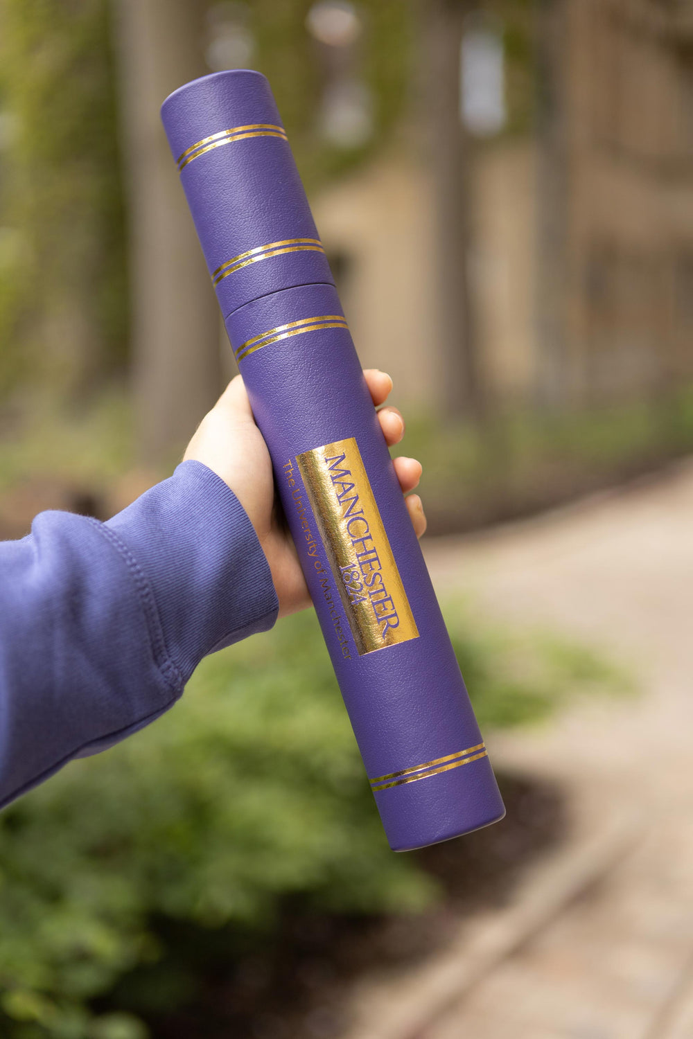 A hand holds a purple diploma tube with gold detailing and "Manchester 1824" text. The background is an outdoor setting with blurred greenery and a path.
