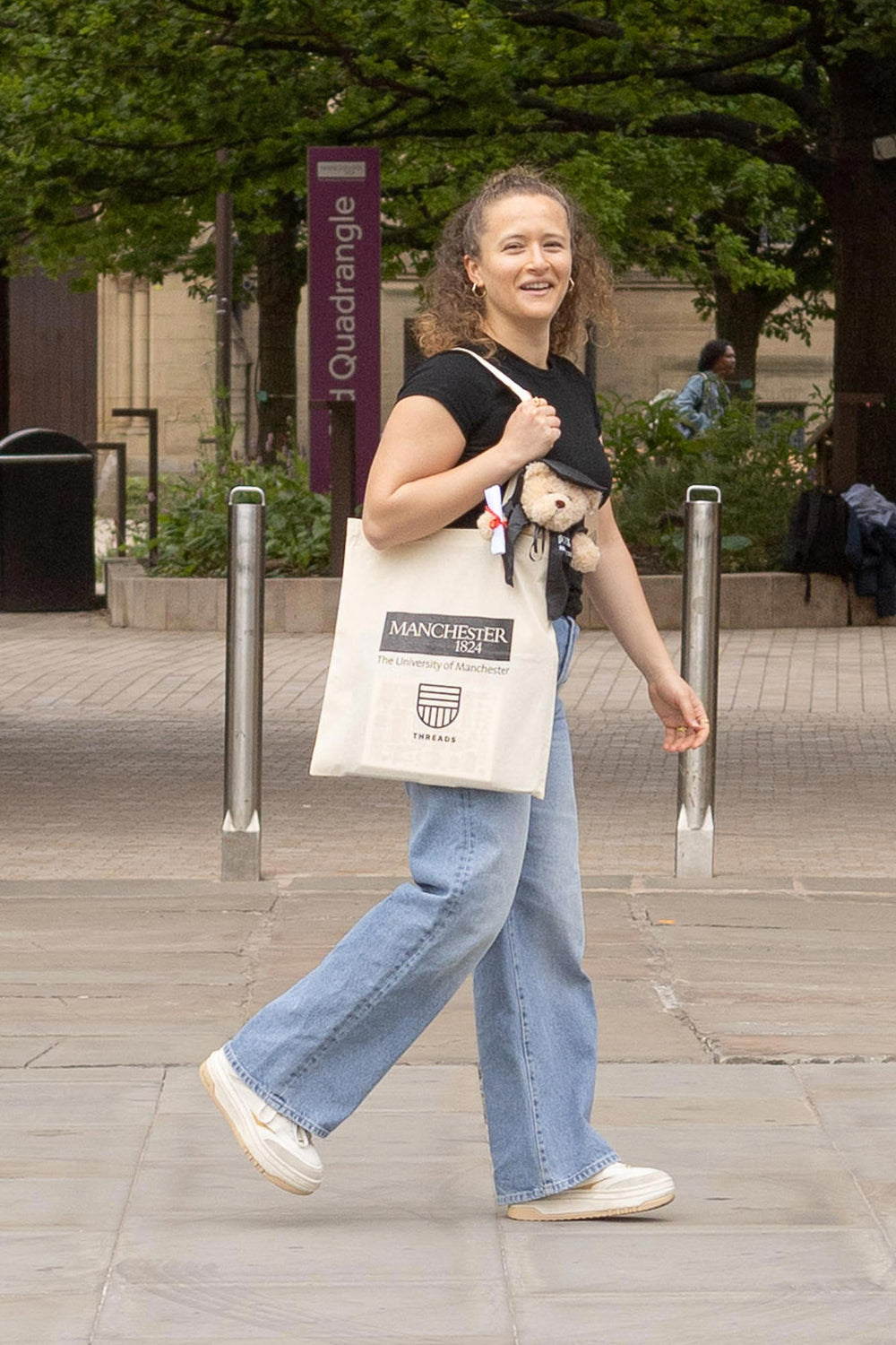 Person walking outside with a smile, carrying a University of Manchester tote bag and a teddy bear. She wears a black top and blue jeans.