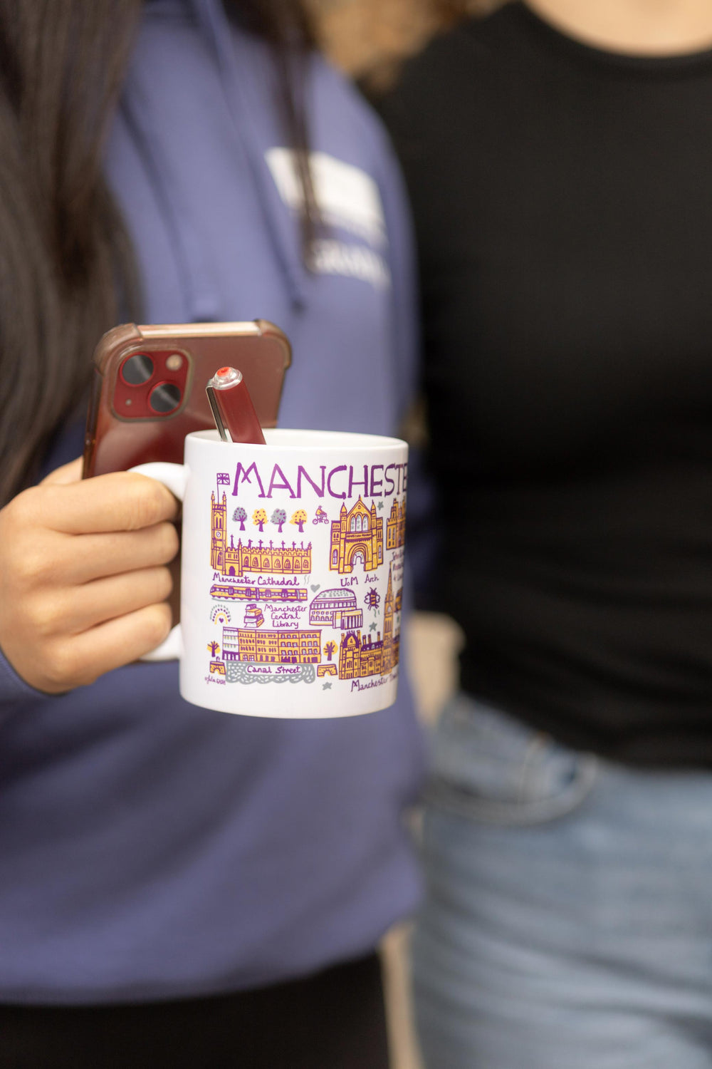 Two people standing side by side. One holds a phone and a white mug with colorful Manchester landmarks. The mood is casual and friendly.